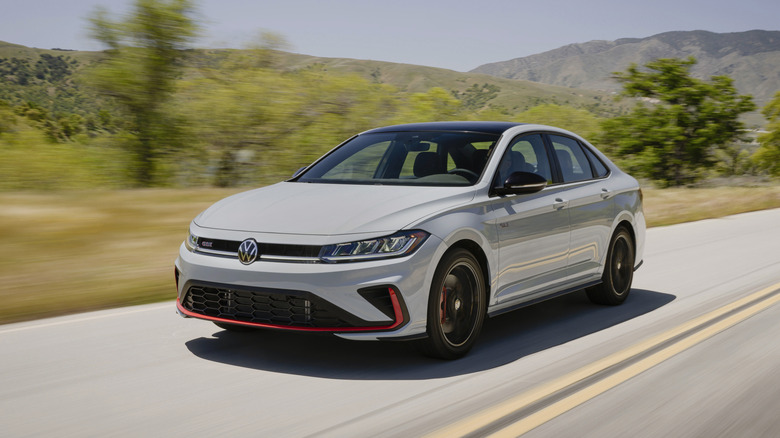 A light gray Volkswagen Jetta GLI sedan with distinctive red front accents driving along a scenic mountain road.