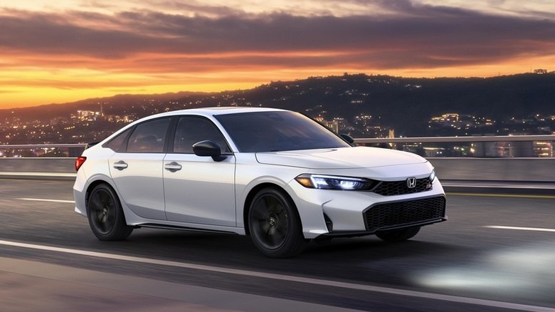 A white 2025 Honda Civic Si sedan driving along a highway bridge at twilight, with city lights illuminating the background hills under a vibrant orange and purple sunset sky.