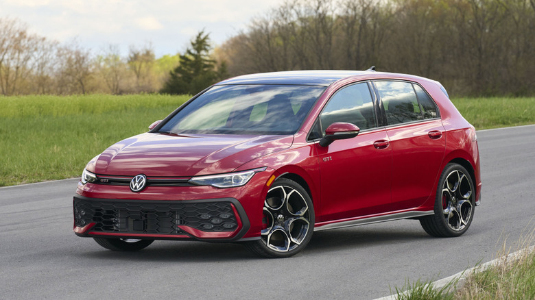 A metallic red 2026 Volkswagen Golf GTI hatchback on an empty two-lane asphalt road bordered by a green field and a line of spring trees.