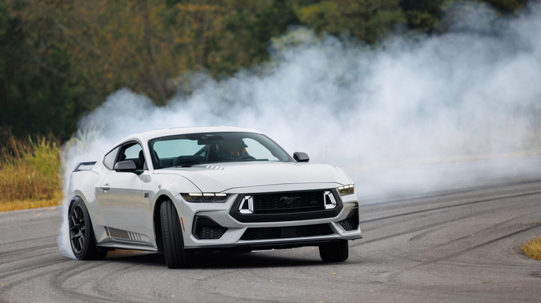 A light gray Ford MustangEcoBoost with an RTR package drifting aggressively on an asphalt track, producing a massive cloud of white smoke.