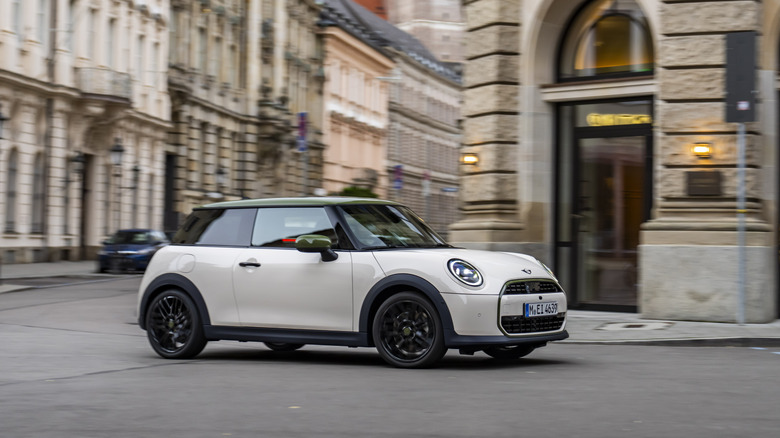 A white Mini Cooper hatchback navigating a street corner with classical architecture.