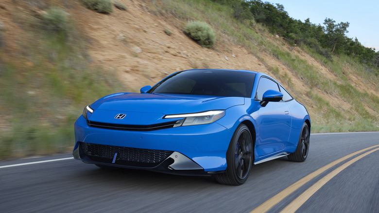 A vibrant blue 2026 Honda Prelude coupe driving through a turn on mountain road flanked by grassy, sunlit hillsides.