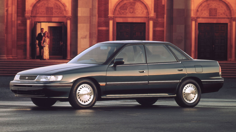 Dark-colored 1990s Subaru Legacy sedan left front angle view parked in front of a fancy building with a couple in formal attire in background