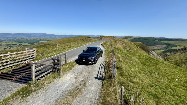 An overhead shot of a Galaxy Purple Pearl WRX parked on a gravel turnout next to a cowcatcher on the peak of a grassy mountain with a view of meadows and blue skies