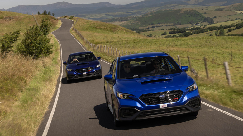 A front shot of a two blue WRXs driving on a rural road surrounded by grass and a view of the green valley below