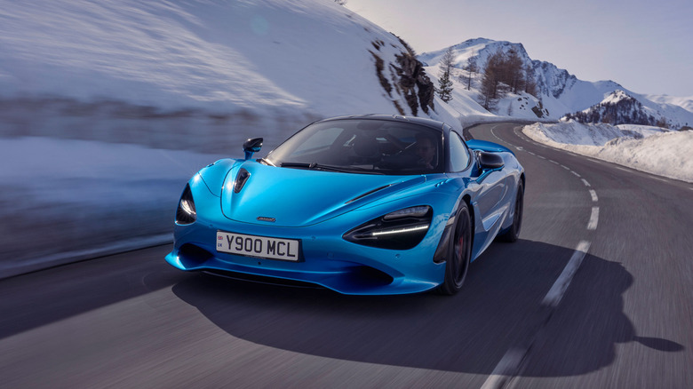 A Ludus Blue 2026 McLaren 750S Coupe driving along a winding, snow-lined mountain road in the French Alps