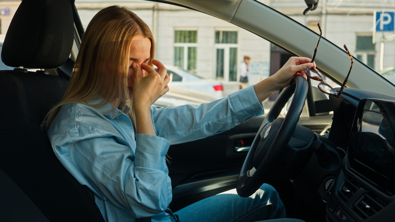 Person sitting a car with their eye glasses off rubbing their nose.