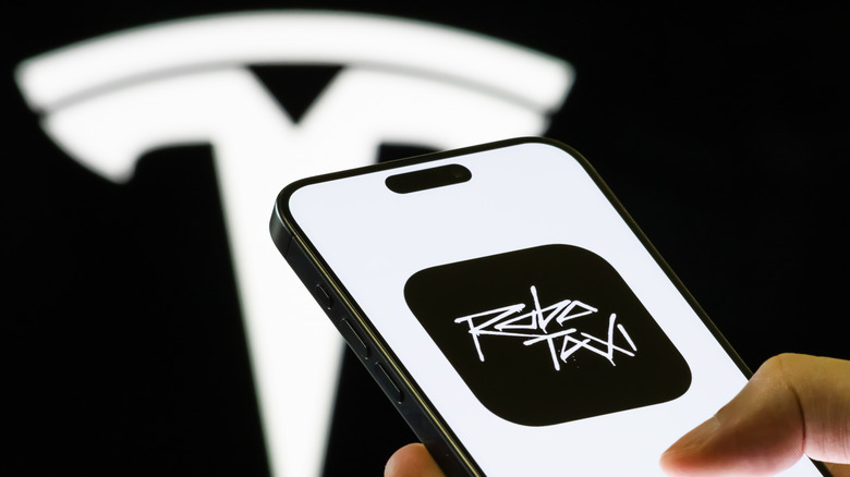 In this photo illustration, a smartphone held in a hand shows the Tesla Robotaxi app, a platform allowing users to join the waitlist for autonomous ride-hailing services using Tesla Model Y vehicles, with the company's branding seen in the background on September 4, 2025 in Chongqing, China.