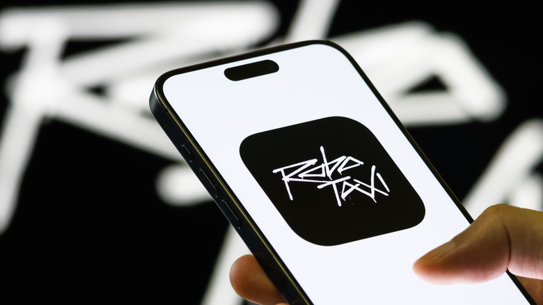 In this photo illustration, a smartphone held in a hand shows the Tesla Robotaxi app, a mobile interface that allows users to join a waitlist for autonomous ride-hailing services using Tesla ModelY vehicles, with the company's branding seen in the background on September 4, 2025
