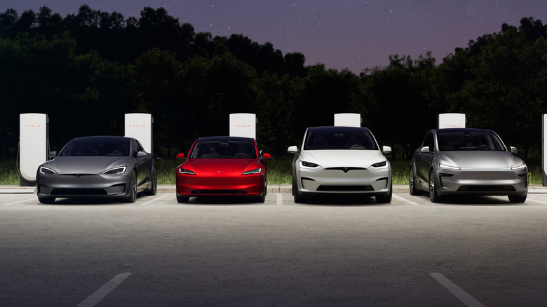 A head-on shot of the four main Tesla models lined up so their names spell S3XY in front of superchargers at dusk