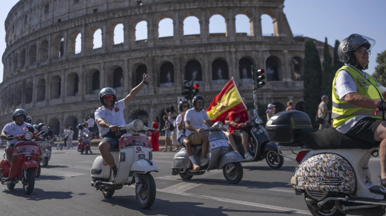 A man in a helmet riding his white Vespa waves to camera, surrounded by fellow Vespa riders as they ride around Rome's Colosseum.
