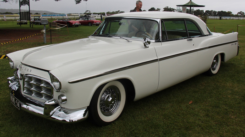 A 1956 cream-colored Chrysler 300B Hardtop parked on grass with a person and other cars in the background