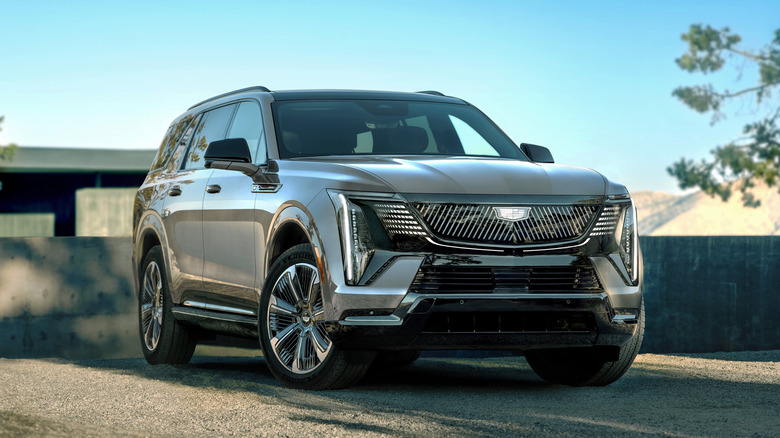A metallic silver 2026 Cadillac Escalade IQL all-electric luxury SUV showcasing its massive illuminated grille and vertical LED headlights while parked on a sunlit gravel driveway.
