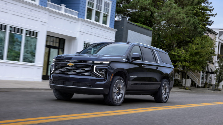 A dark blue Chevrolet Suburban High Country extended wheelbase SUV driving through a town street past a modern blue and white building.