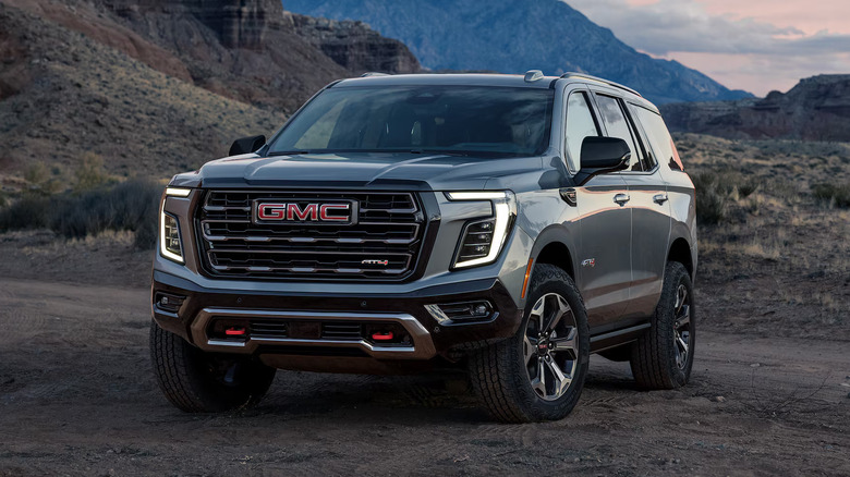 A front 3/4 view of a silver-grey GMC Yukon AT4 Ultimate full-size off-road SUV parked on a dirt trail amidst a rugged desert canyon landscape with red rock cliffs.