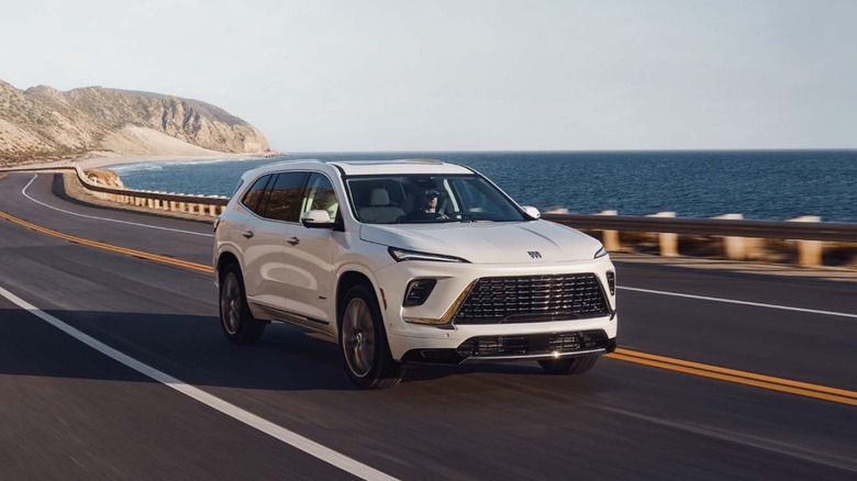 A white Buick Enclave three-row crossover cruising along a scenic two-lane coastal highway with the ocean and steep rocky cliffs in the background.