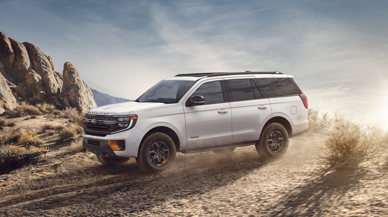A star white metallic 2026 Ford Expedition Tremor full-size off-road SUV creates dust as it drives over a rugged, sunlit dirt trail bordered by massive boulders.