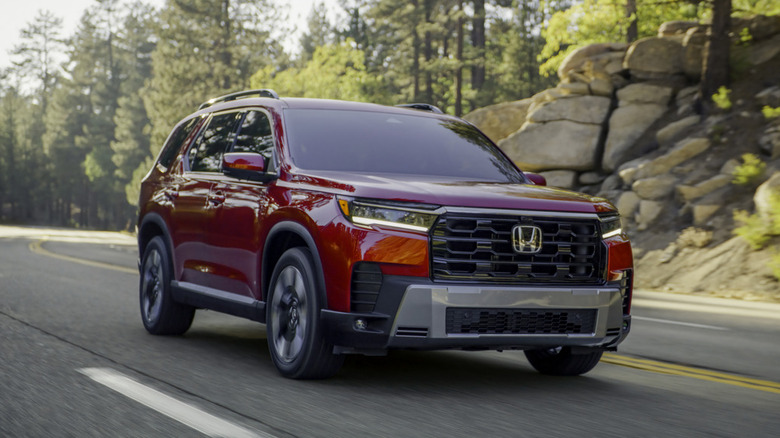 A red 2026 Honda Pilot Elite mid-size SUV driving along a winding mountain road lined with pine trees and sunny rock formations.