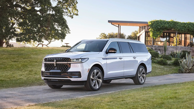 A white Lincoln Navigator L extended luxury SUV parked on a paved path in front of a modern glass-and-wood house with a green lawn.