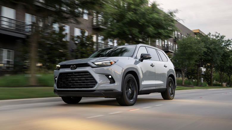 A grey Toyota Grand Highlander Nightshade edition with black wheels driving past dark brick residential buildings with a motion blur effect.