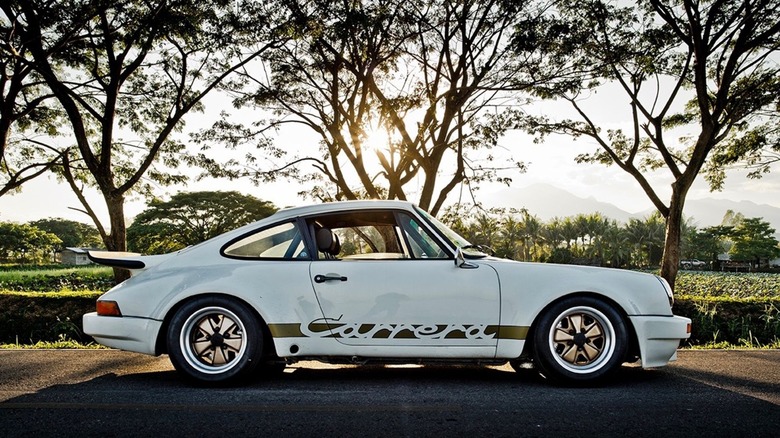 A side shot of a white Porsche Carrera RSR 3.0 Replica parked in front of several trees, a field, and a low sun