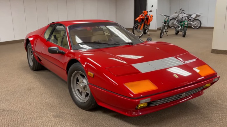A red 1976 Ferrari 512 BB parked in an indoor showroom with several dirt bikes in the background