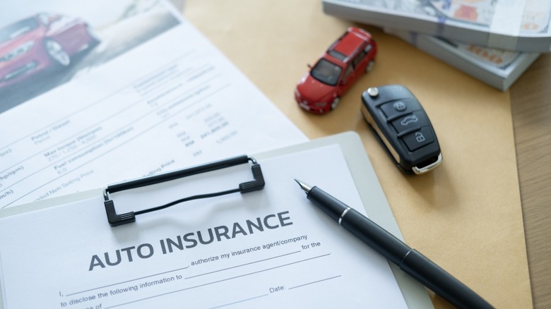 A paper that reads "Auto Insurance" on a clipboard placed on a desk with a pen, toy car, and key fob