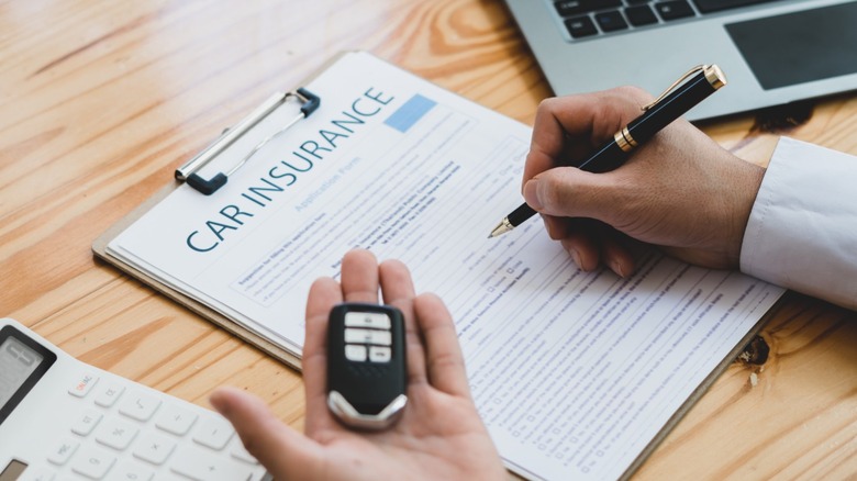 A hand holds a key fob while another hand signs a car insurance document