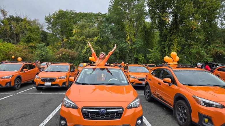 A woman dressed in orange posing in the sunroof of a Sunshine Orange Subaru Crosstrek among many other identical cars