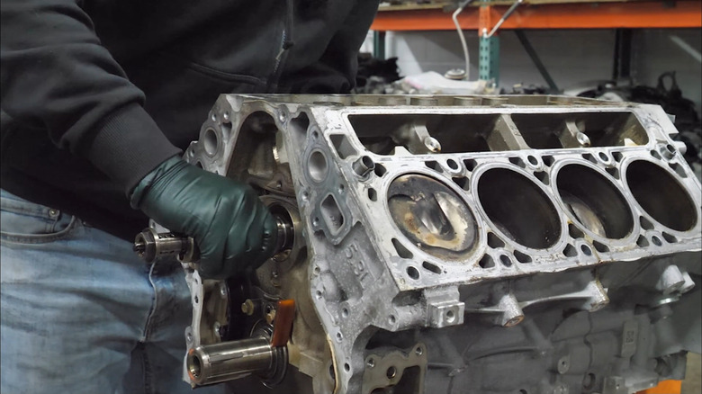 A man in green gloves, blue jeans, and a black sweatshirt pulling the camshaft out of a GM 5.3-liter V8 missing its heads
