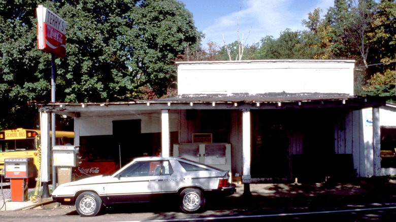 A 1980s Dodge Charger 2.2 parked at a rural gas station, with trees in the background.