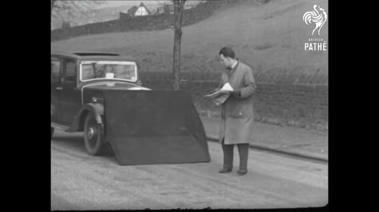 A pedestrian in the 1930s, holds his paper and looks towards the car with the pedestrian catcher extended as it approaches to scoop up said pedestrian in the road.