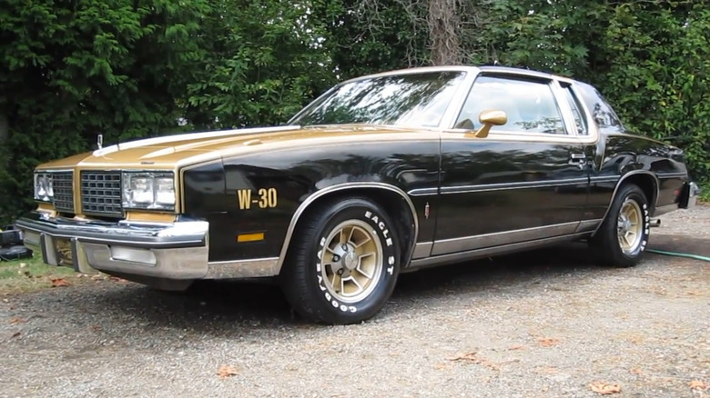 A black and gold 1980 Oldsmobile 442 W-30 sits on a gravel road in front of trees