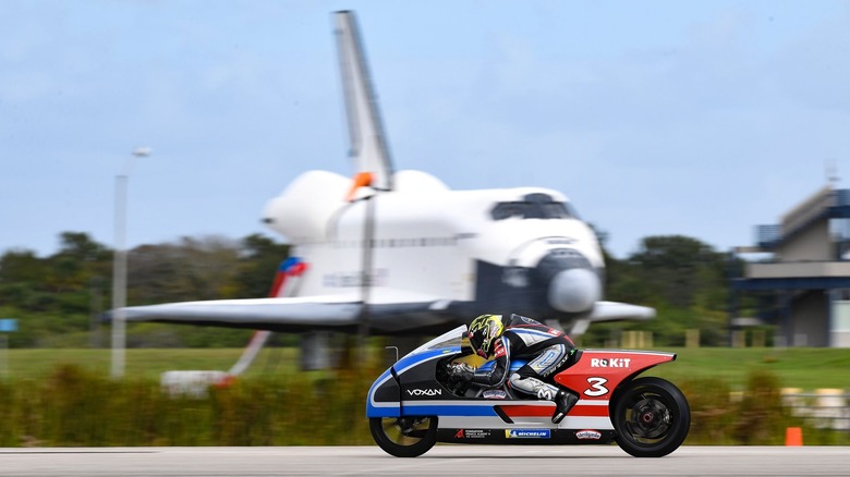 A running shot of the Voxan Wattman electric motorcycle on a runway in front of the space shuttle.