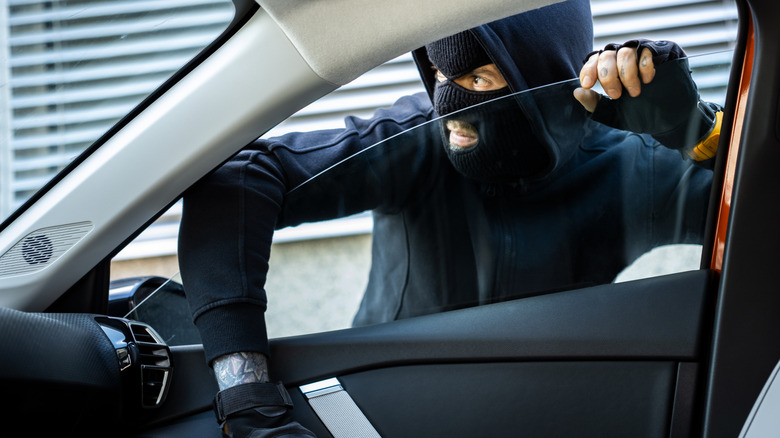 A criminal in a balaclava and dark clothes trying to break into a car through an open window.
