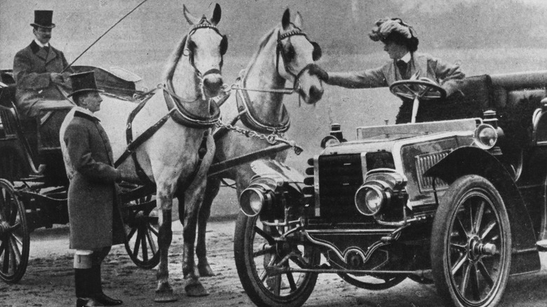 A 1904 photograph of a set of horses next to a motorcar, with one horse being pet by the car's driver