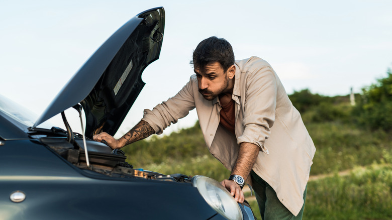 A worried man looking at the engine of a dark car with the hood raised