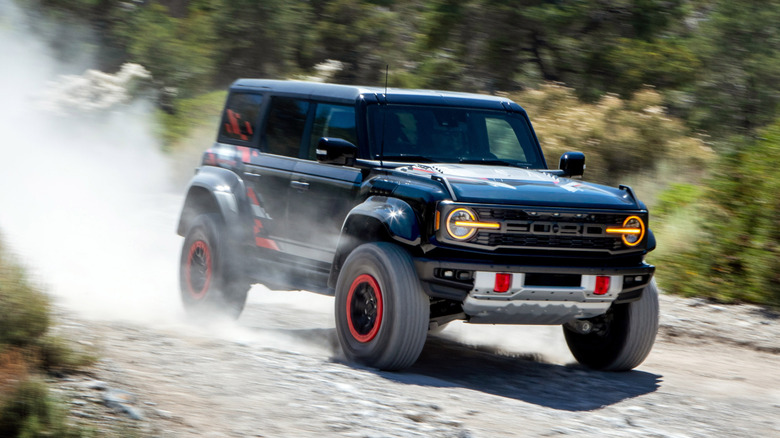 Front three quarters shot of a black Bronco Raptor Code Orange kicking up dust while driving over gravel past green bushes