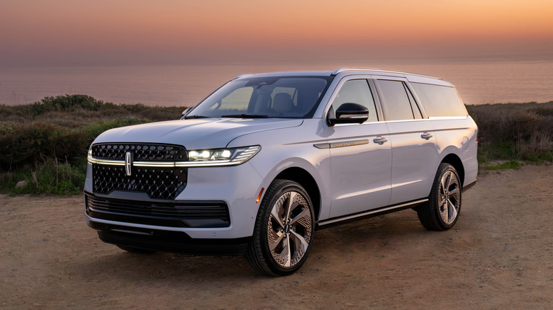 A front three quarters shot of a white Lincoln Navigator L parked on dirt in front of shrubs and the ocean at dusk