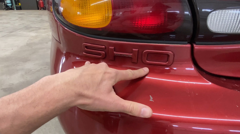 A man's hand shows the badge on the back end of a red 1997 Ford Taurus SHO with the 3.4-liter Yamaha 60-degree V8
