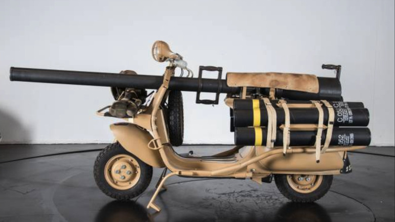 A sand-colored Vespa 150 TAP equipped with a gun, in a studio with a white wall and black round table.