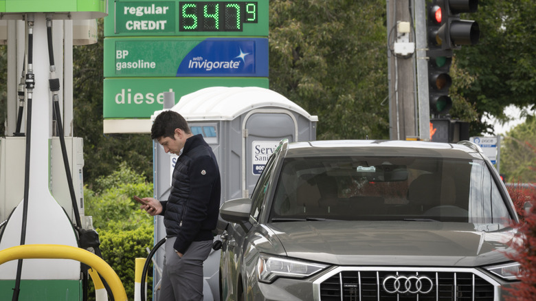 A driver using a cellphone while fueling up a gray Audi SUV