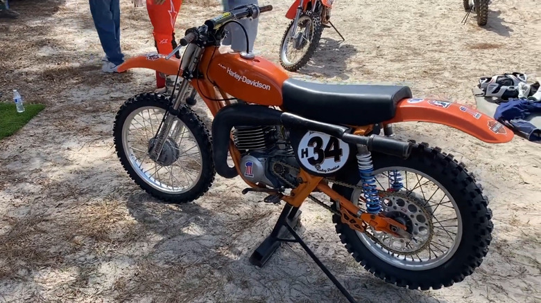 An orange Harley-Davidson MX250 dirt bike at a North Florida motocross park, parked on dirt with people's legs and other bikes visible in the background