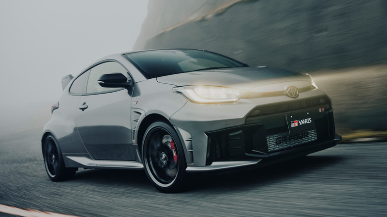 A dramatic low-angle motion shot of a grey Toyota GR Yaris all-wheel-drive hot hatch driving through mist along a cliffside road.