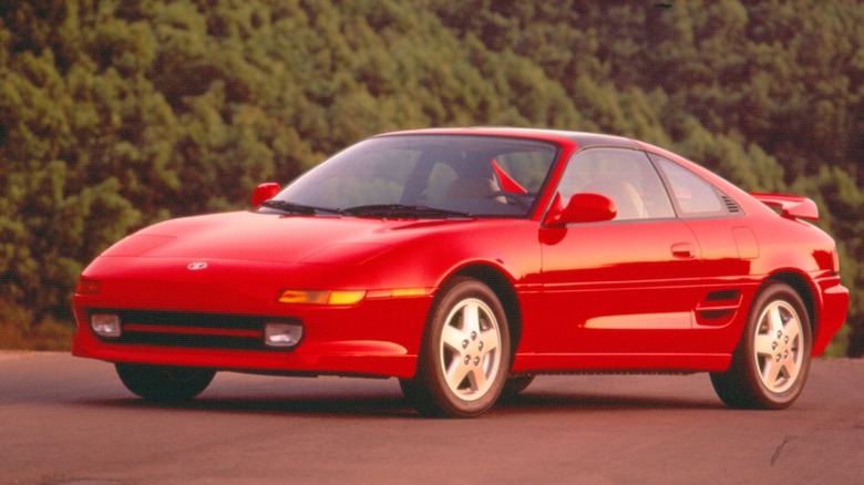 A red 1994 Toyota MR2 Turbo mid-engine sports coupe parked on tarmac with lush green foliage in the background, front three-quarter view.