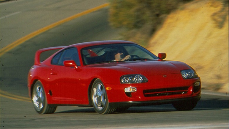 A red 1998 Toyota Supra Turbo Mk4 coupe driving on a canyon road, front three-quarter view.