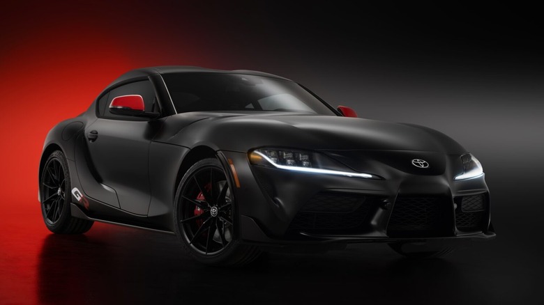 A matte black 2026 Toyota GR Supra Final Edition featuring red mirror caps and illuminated LED DRLs against a dramatic red and black background.