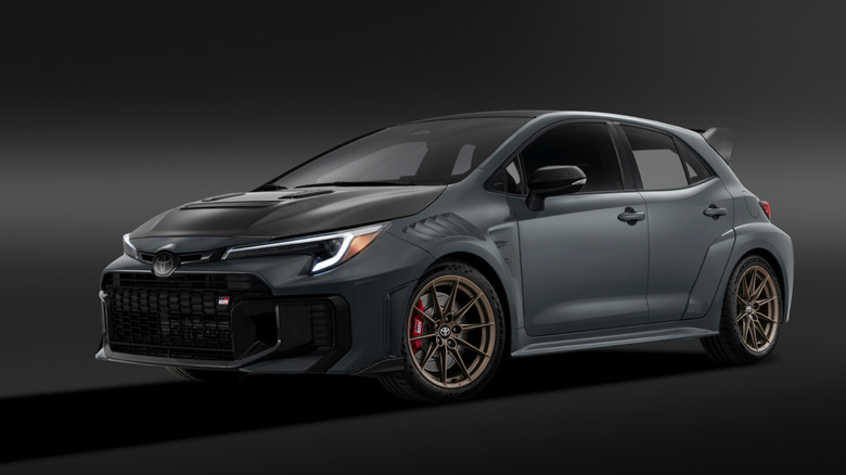 A aggressive matte dark grey Toyota GRMN Corolla featuring a vented carbon fiber hood, fender louvers, bronze wheels, and carbon rear wing in a dark studio setting.