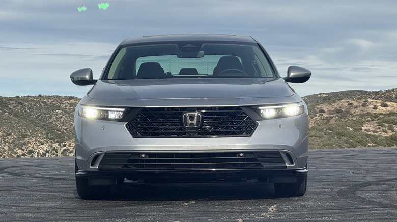 A head-on shot of the silver Accord Hybrid Touring parked on asphalt in front of a shrub-covered hill and a cloudy sky