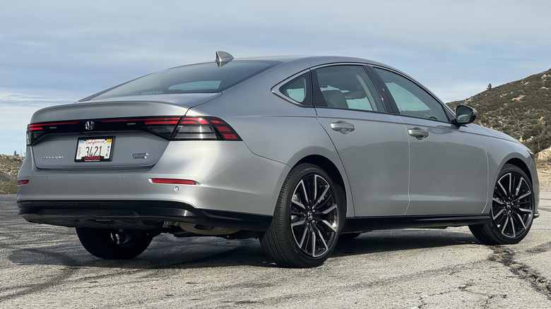 A rear three-quarters shot of the silver Accord Hybrid Touring parked on asphalt in front of a cloudy sky and a shrub-covered hill